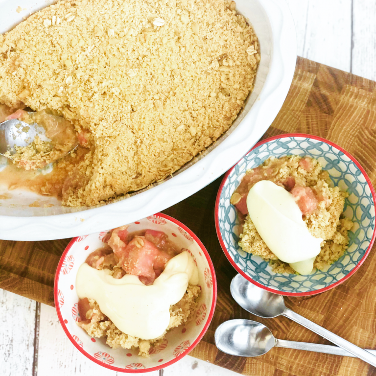 homemade rhubarb crumble being spooned into two serving bowls - rhubarb crumble has rich vanilla custard drizzled on top