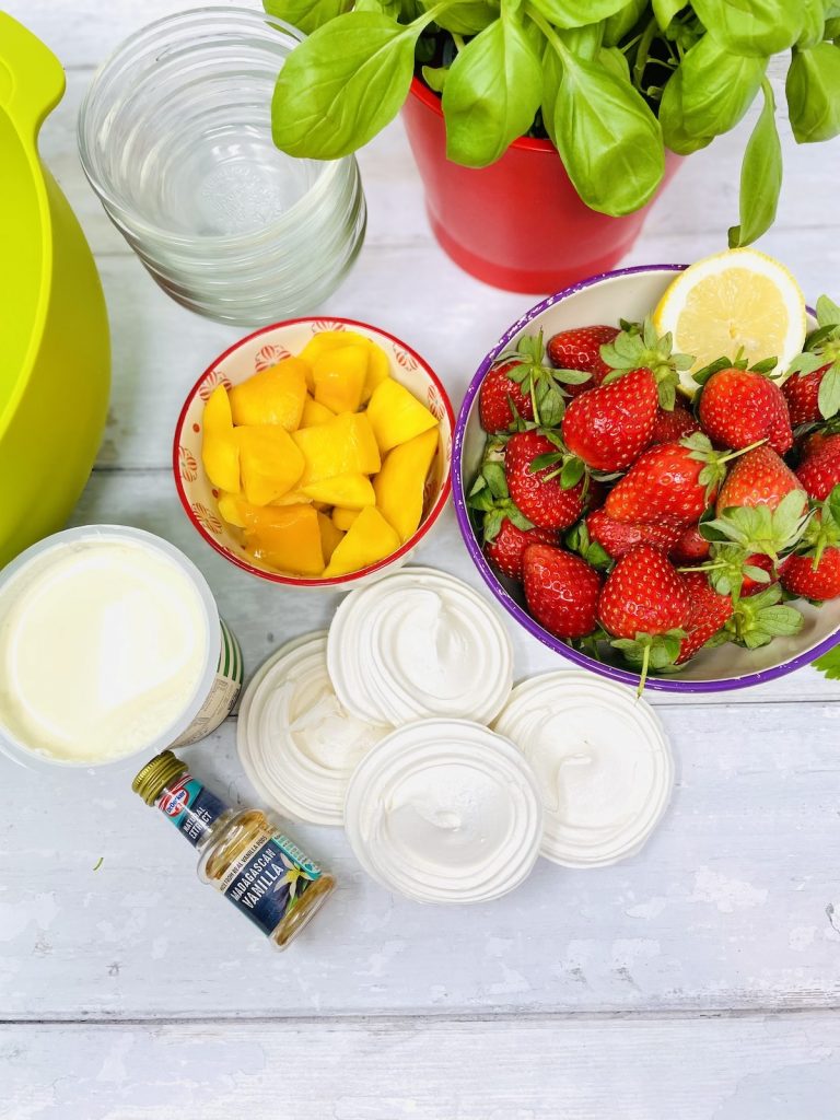 Eton mess ingredients on a white table - strawberries, cream, meringue nests, vanilla extract and mango chunks