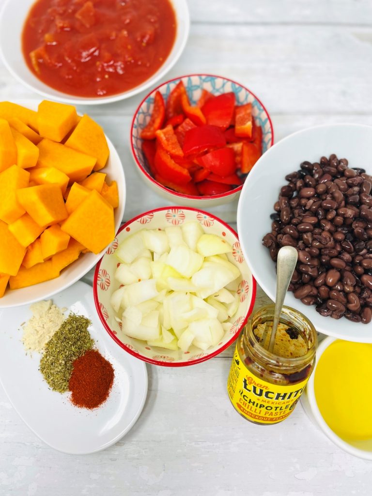 ingredients for butternut squash on a white table - butternut squash cubes, red pepper, black beans, chopped onions, canned tomatoes, chipotle chilli paste, garlic powder, dried oregano, smoked paprika