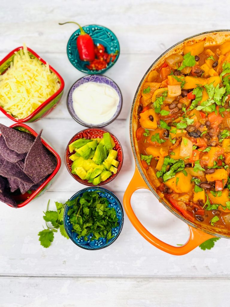 butternut squash chilli veggie chilli recipe served with side dishes of coriander, avocado chunks, yogurt, sliced chillies, blue corn tortillas and grated cheese