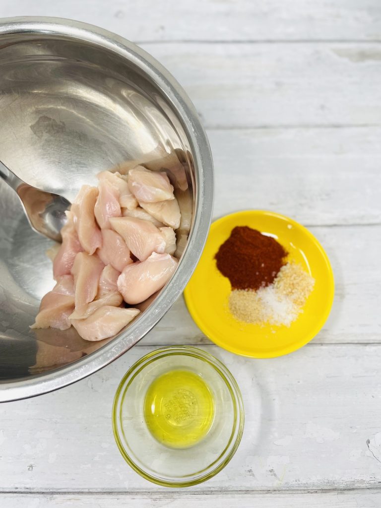 ingredients for paprika chicken on a table - includes raw chicken breast pieces, jar of olive oil, yellow saucer with paprika, garlic powder, onion granules and pinch of salt