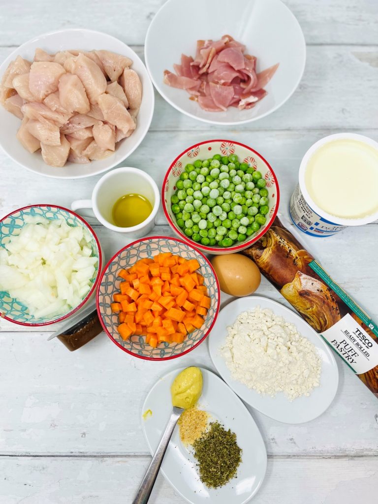 ingredients for chicken and bacon pie on a white table top - they include diced chicken breast, chopped bacon, frozen peas, diced carrots and onions, small saucer with herbs and spices, saucer of plain flour, pot of cream and sheet of puff pastry in a packet