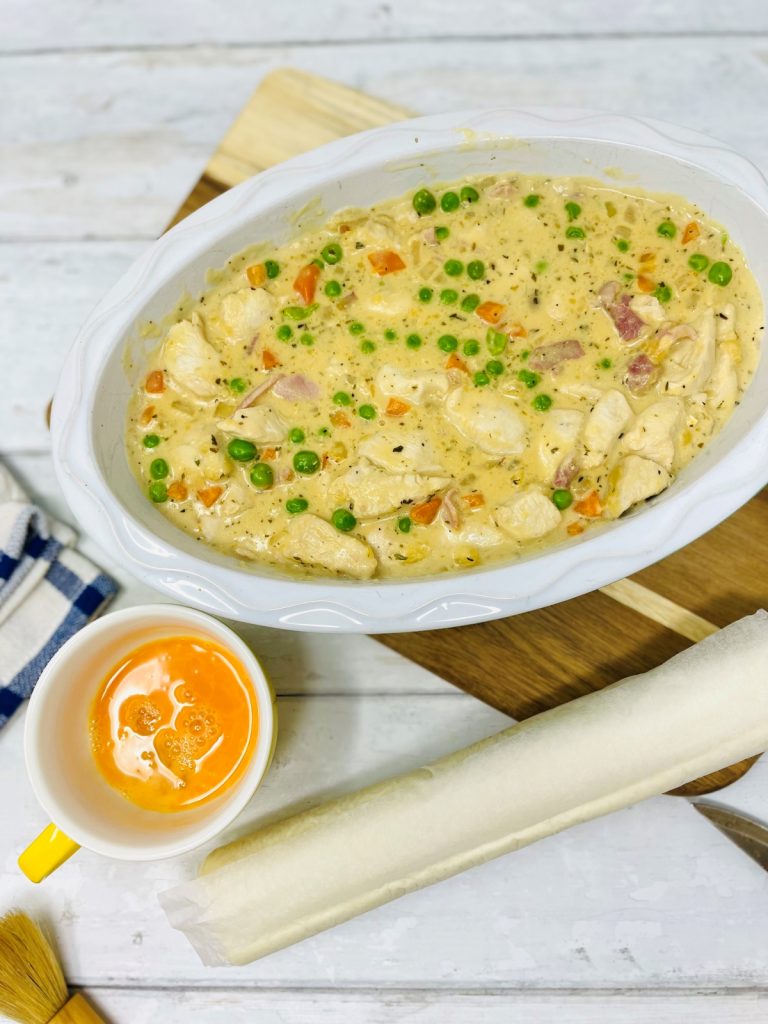 creamy chicken and bacon pie filling tipped into a pie dish. Cup of beaten egg and sheet or rolled up puff pastry on counter top next to pie dish