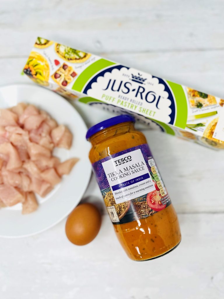 chicken tikka pasty ingredients on a white table - diced chicken breast on a plate, box of jus-roll puff pastry sheet, jar of Tesco tikka masala sauce and one egg