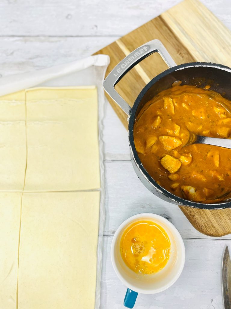 puff pastry sheet cut into four sections, pan of chicken tikka masala next to the pastry, small bowl of beaten egg by the pan