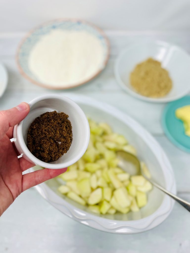 chopped pears in an ovenproof dish - small bowl of brown sugar being held over the dish waiting to be tipped in. Bowl of flour, brown sugar and a saucer of butter in the background